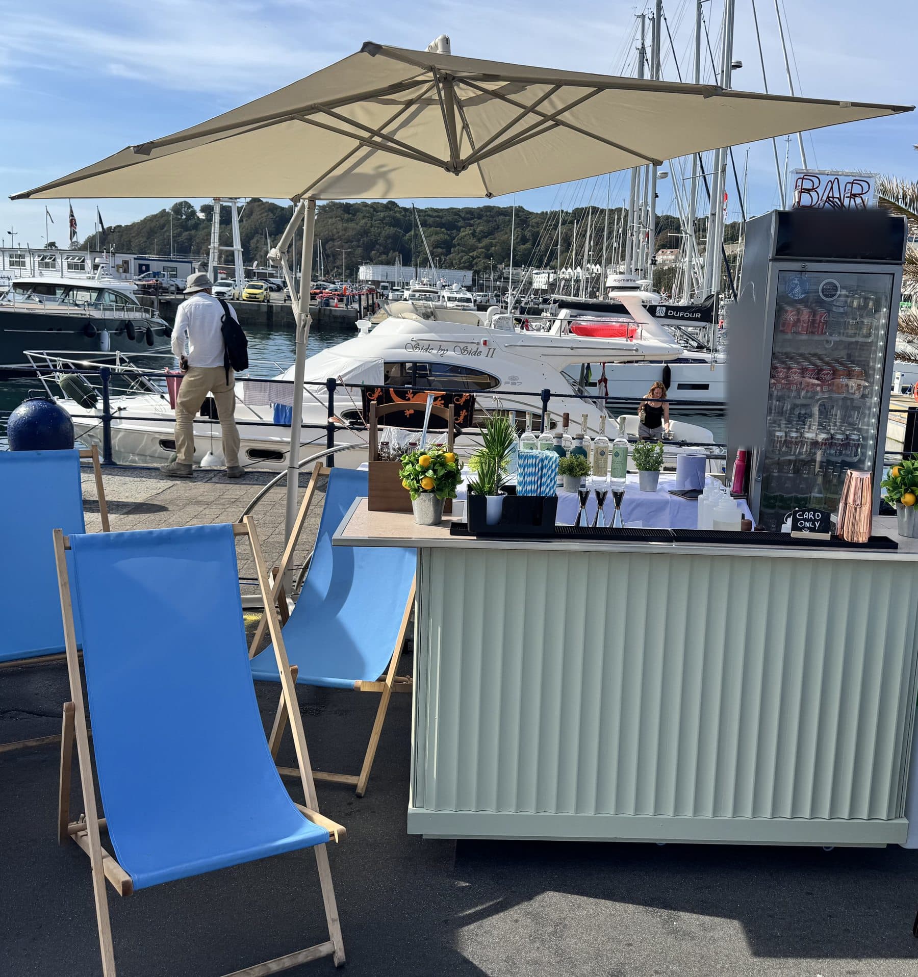 The Dirty Olive bar under a parasol on St Peter Port harbour, with blue deckchairs and yachts behind