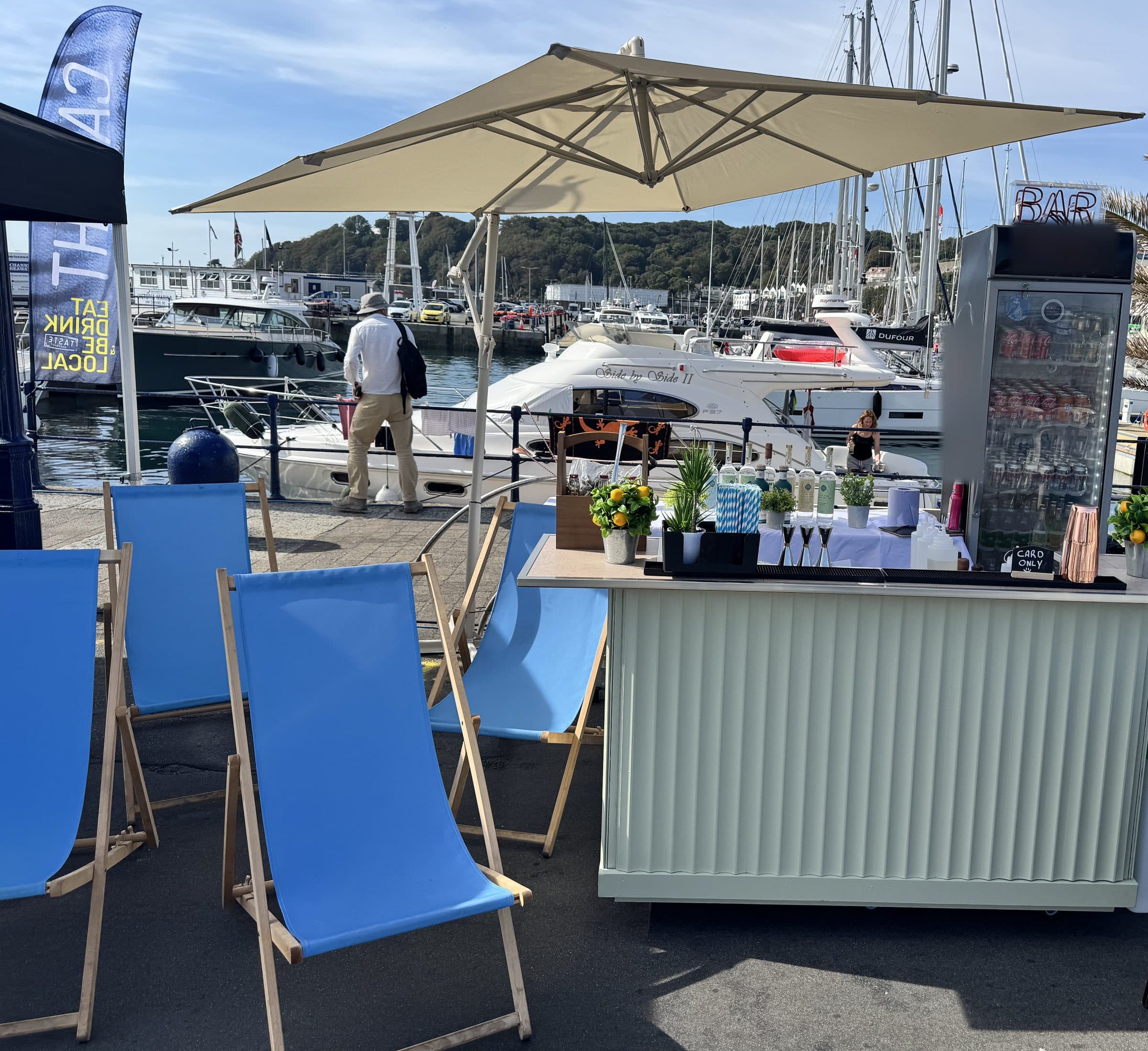 The Dirty Olive bar under a parasol on St Peter Port harbour, blue deckchairs in front and yachts moored behind