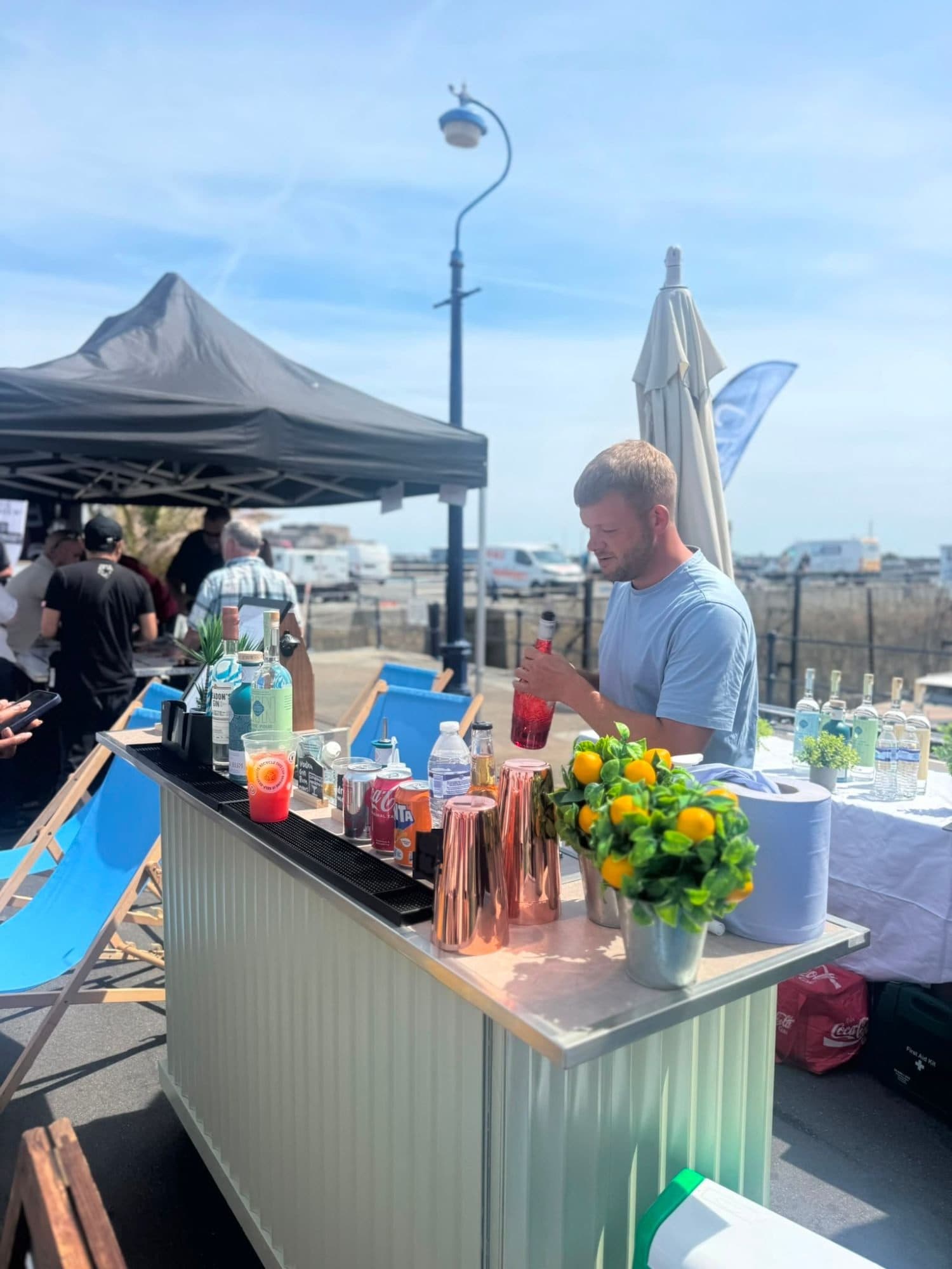 A bartender pouring a drink at the green Dirty Olive mobile bar beside a Guernsey harbour